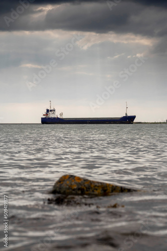 Cargo ship in focus. Rough stone rocks in foreground out of focus. Danger of the ocean. World goods shipping industry. Export and import business. Dark and moody sky and water surface. © mark_gusev