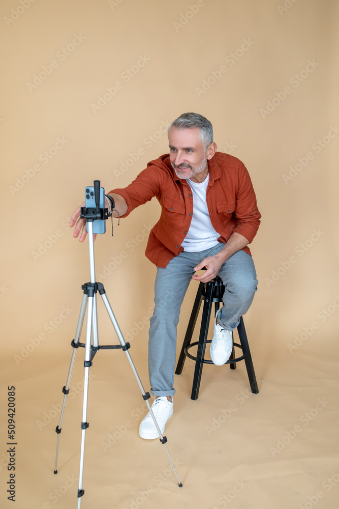 A man in a red jacket sitting on a stool in a studio and making selfie