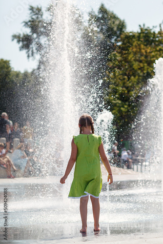 A young girl in a light green dress barefoot dances by a fountain in the city center.