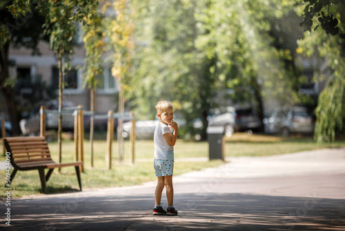 A little boy is standing in the shade of trees in a city park, the child is alone he is afraid of getting lost