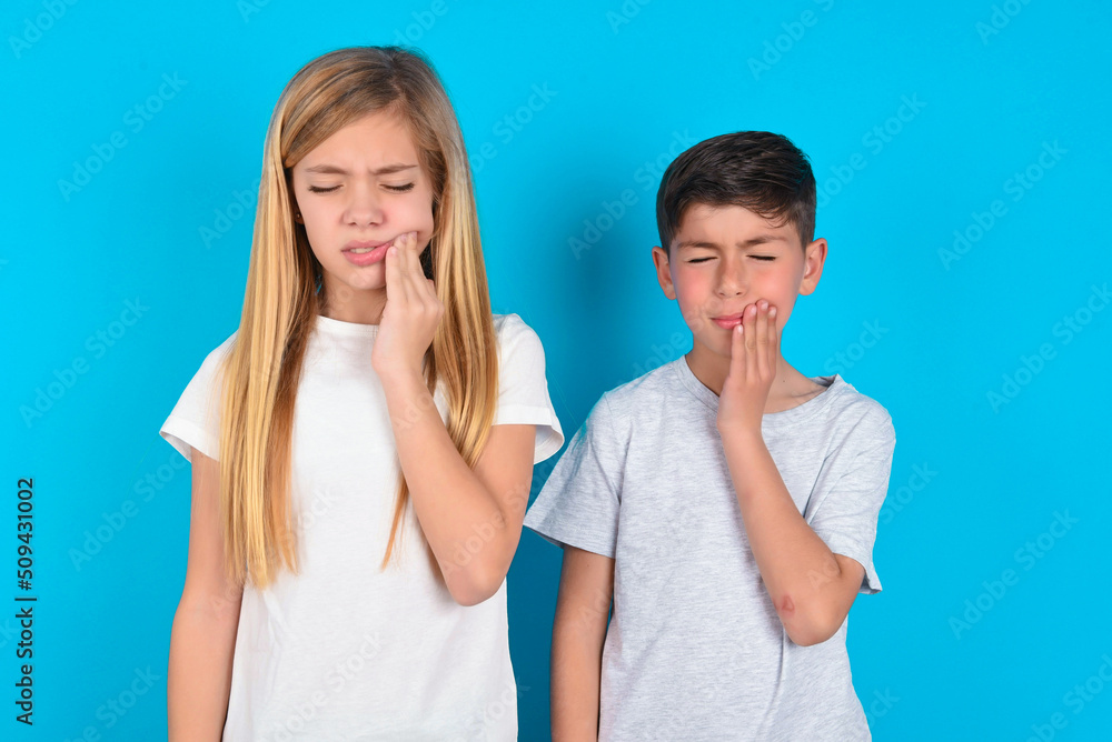 Tooth ache concept. two kids boy and girl standing over blue studio background feeling pain, holding his cheek with hand, suffering from bad toothache, looking at camera with painful expression Tooth ache concept. two kids boy and girl standing over blue studio background feeling pain, holding his cheek with hand, suffering from bad toothache, looking at camera with painful expression