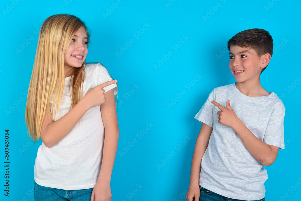Happy cheerful smiling two kids boy and girl standing over blue studio background looking and pointing aside with hand. Copy space and advertisement concept. Happy cheerful smiling two kids boy and girl standing over blue studio background looking and pointing aside with hand. Copy space and advertisement concept.
