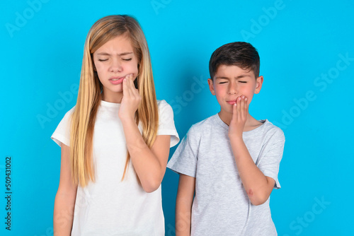 Tooth ache concept. two kids boy and girl standing over blue studio background feeling pain, holding his cheek with hand, suffering from bad toothache, looking at camera with painful expression