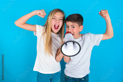two kids boy and girl standing over blue studio background communicates shouting loud holding a megaphone, expressing success and positive concept, idea for marketing or sales.