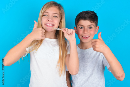 two kids boy and girl standing over blue studio background holding an invisible braces aligner and rising thumb up, recommending this new treatment. Dental healthcare concept.