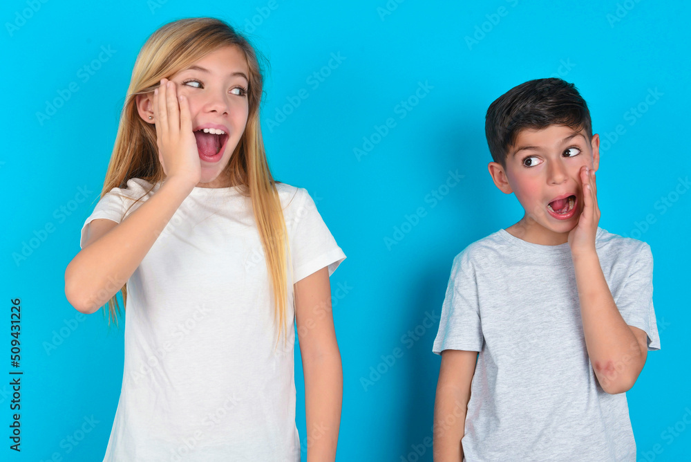 two kids boy and girl standing over blue studio background excited looking to the side hand on face. Advertisement and amazement concept. two kids boy and girl standing over blue studio background excited looking to the side hand on face. Advertisement and amazement concept.