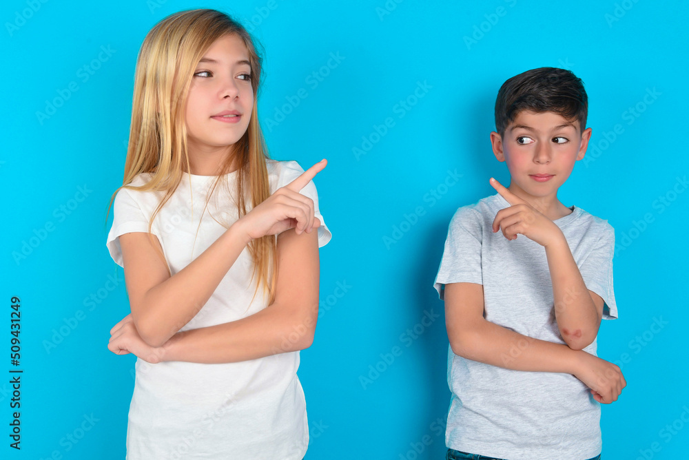 Portrait of two kids boy and girl standing over blue studio background posing on camera with tricky look, presenting product with index finger. Advertisement concept. Portrait of two kids boy and girl standing over blue studio background posing on camera with tricky look, presenting product with index finger. Advertisement concept.