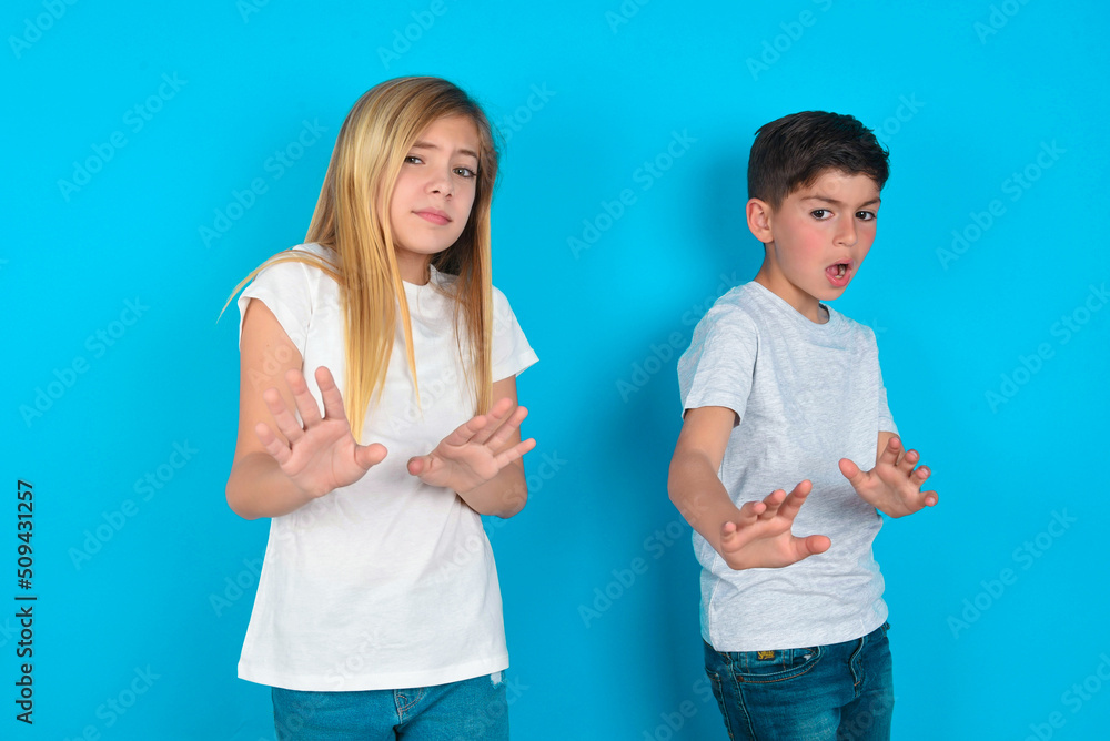 Afraid two kids boy and girl standing over blue studio background, makes terrified expression and stop gesture with both hands saying: Stay there. Panic concept. Afraid two kids boy and girl standing over blue studio background, makes terrified expression and stop gesture with both hands saying: Stay there. Panic concept.