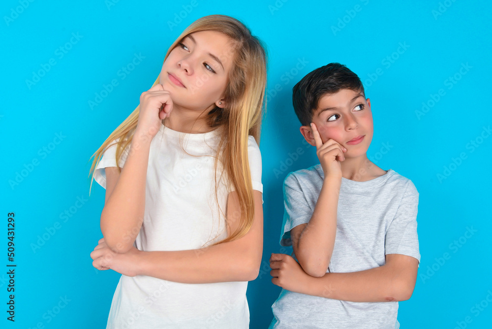 Face expressions and emotions. Thoughtful two kids boy and girl standing over blue studio background holding hand under his head, having doubtful look. Face expressions and emotions. Thoughtful two kids boy and girl standing over blue studio background holding hand under his head, having doubtful look.