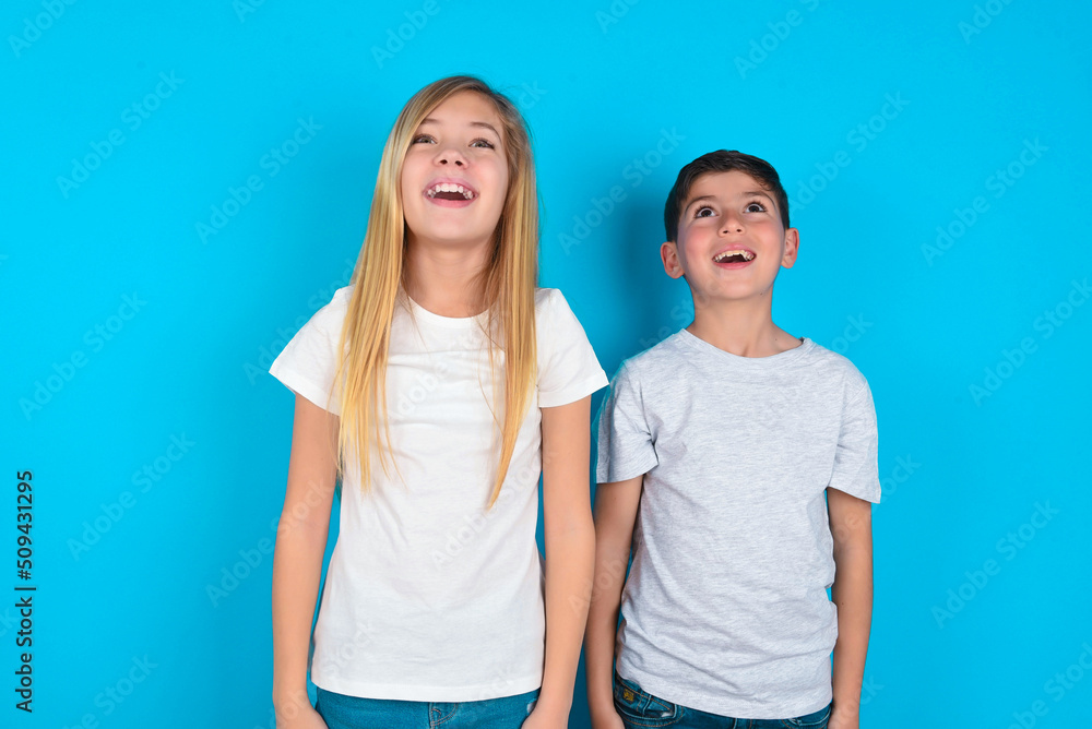 Surprised two kids boy and girl standing over blue studio background, shrugs shoulders, looking sideways, being happy and excited. Sudden reactions concept. Surprised two kids boy and girl standing over blue studio background, shrugs shoulders, looking sideways, being happy and excited. Sudden reactions concept.