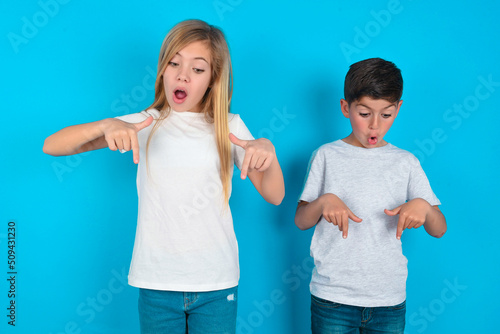 Amazed two kids boy and girl standing over blue studio background points down with fore fingers, opens mouth being shocked. Advertisement concept. photo