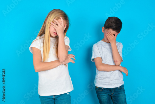 two kids boy and girl standing over blue studio background making facepalm gesture while smiling amazed with stupid situation. photo