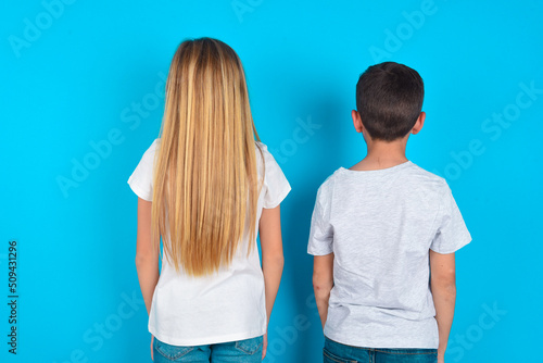 two kids boy and girl standing over blue studio background standing backwards looking away with arms on body. photo