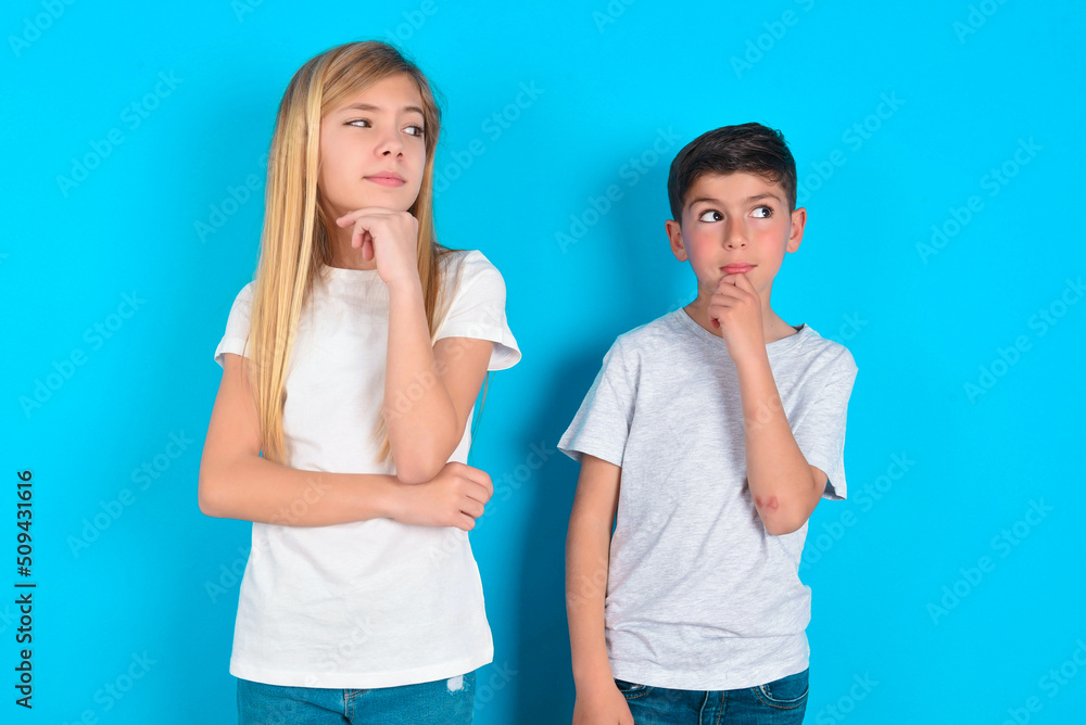 Shot of contemplative thoughtful two kids boy and girl standing over blue studio background keeps hand under chin, looks thoughtfully upwards. Shot of contemplative thoughtful two kids boy and girl standing over blue studio background keeps hand under chin, looks thoughtfully upwards.
