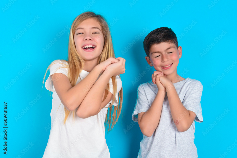 Dreamy two kids boy and girl standing over blue studio background with pleasant expression, closes eyes, keeps hands crossed near face, thinks about something pleasant Dreamy two kids boy and girl standing over blue studio background with pleasant expression, closes eyes, keeps hands crossed near face, thinks about something pleasant