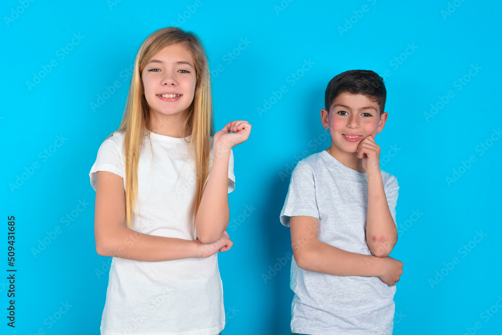 Cheerful two kids boy and girl standing over blue studio background with hand near face. Looking with glad expression at the camera after listening to good news. Confidence. Cheerful two kids boy and girl standing over blue studio background with hand near face. Looking with glad expression at the camera after listening to good news. Confidence.
