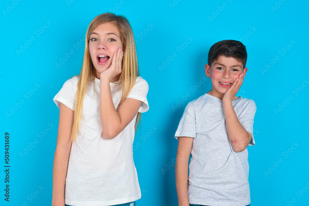 Shocked, astonished two kids boy and girl standing over blue studio backgroun looking surprised in full disbelief wide open mouth with hand near face. Positive emotion facial expression body language. Shocked, astonished two kids boy and girl standing over blue studio backgroun looking surprised in full disbelief wide open mouth with hand near face. Positive emotion facial expression body language.