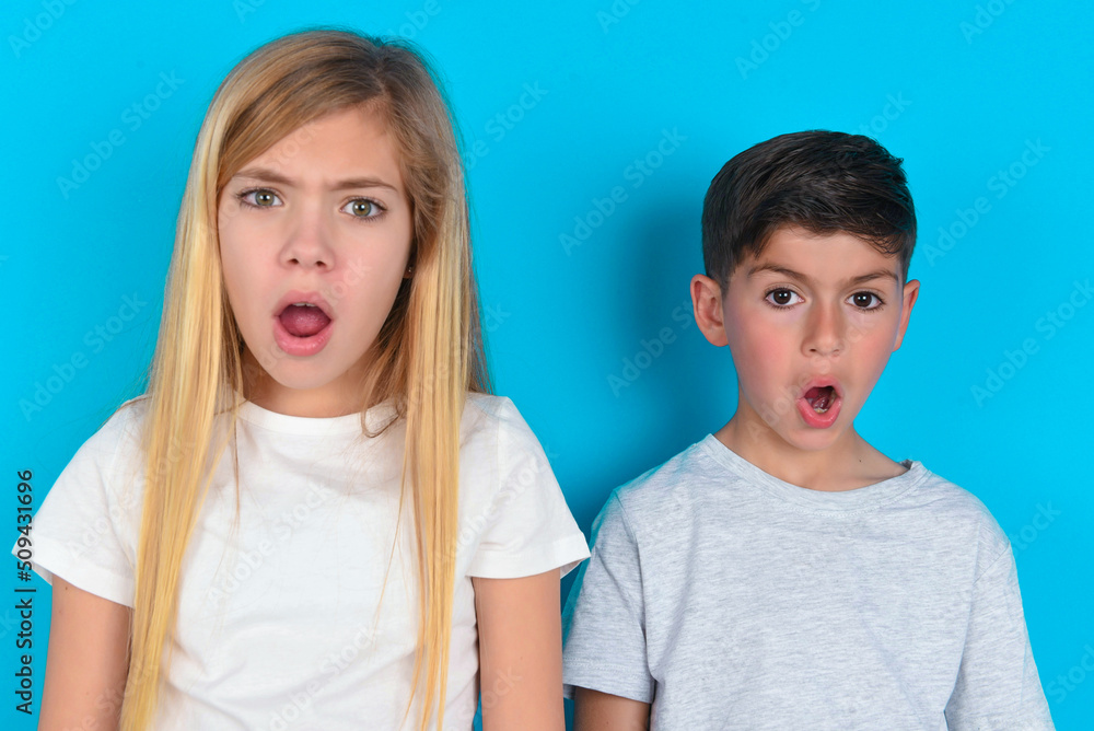 Expressive facial expressions. Shocked stupefied two kids boy and girl standing over blue studio background, keeps jaw dropped feels stunned from what he sees aside. Expressive facial expressions. Shocked stupefied two kids boy and girl standing over blue studio background, keeps jaw dropped feels stunned from what he sees aside.