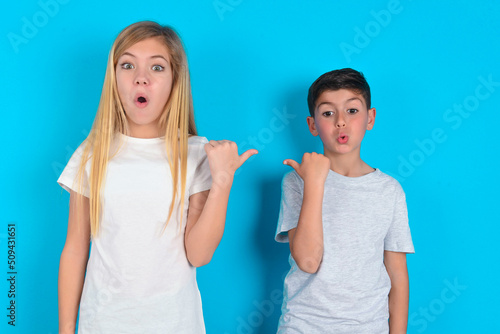 Shocked two kids boy and girl standing over blue studio background points with thumb away, indicates something. Check this out. Advertisement concept.