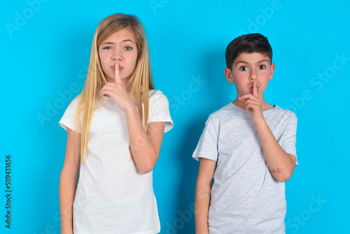 two kids boy and girl standing over blue studio background makes hush gesture, asks be quiet. Don't tell my secret or not speak too loud, please!