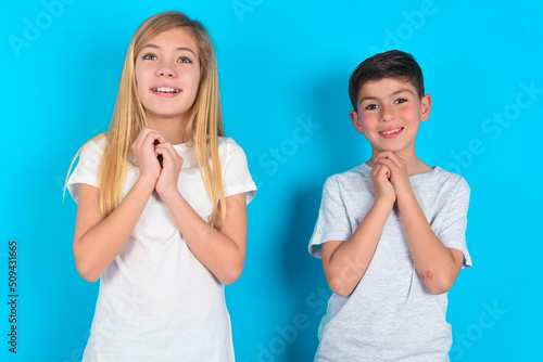 Dreamy charming two kids boy and girl standing over blue studio background with pleasant expression, keeps hands crossed near face, excited about something pleasant.