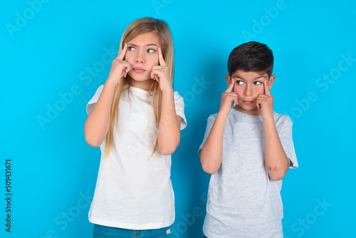 two kids boy and girl standing over blue studio background with thoughtful expression, looks away, keeps hand near face, thinks about something pleasant.