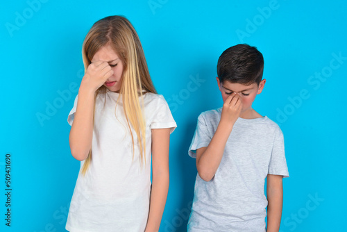 Very upset, two kids boy and girl standing over blue studio background touching nose between closed eyes, wants to cry, having stressful relationship or having troubles with work