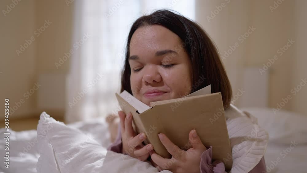 Young happy little woman reading book lying in cozy bed smiling looking away. Portrait of charming little person dreaming enjoying hobby on weekend at home Young happy little woman reading book lying in cozy bed smiling looking away. Portrait of charming little person dreaming enjoying hobby on weekend at home