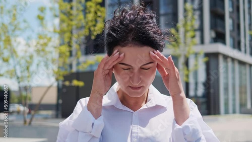 Tired exhausted young adult businesswoman, office worker having headache standing outdoors. Stressed overworked female massages head. Woman feeling terrible pain, migraine headaches, weather changes.