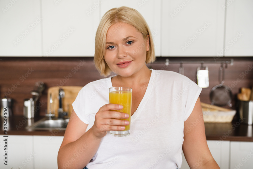 Cheerful beautiful plus size plump woman drinking healthy orange fruit juice for vitamins at home kitchen, having breakfast looking at camera Cheerful beautiful plus size plump woman drinking healthy orange fruit juice for vitamins at home kitchen, having breakfast looking at camera