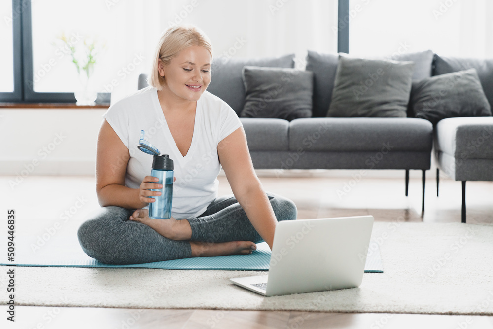 Caucasian young woman plus size plump athlete in fitness clothes drinking water after yoga training, slimmimg shaping on online video tutorial laptop at home Caucasian young woman plus size plump athlete in fitness clothes drinking water after yoga training, slimmimg shaping on online video tutorial laptop at home