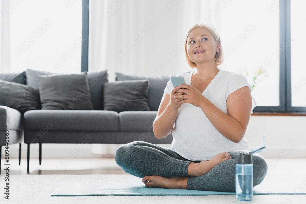 Dreamy caucasian young athlete plus size plump woman in sporty clothes using online mobile application for fitness training, counting burning calories, relaxing on mat at home