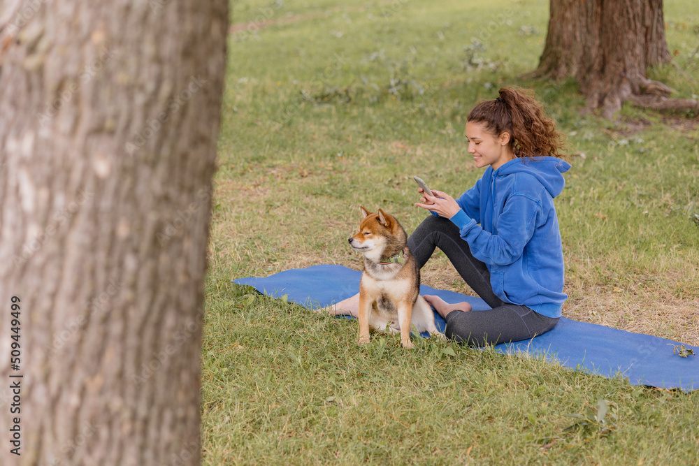 a woman with a dog in the park does sports or fitness on a yoga mat. a European woman with curly hair in a blue hoodie is chatting on a smartphone and playing with a dog. a happy woman is resting a woman with a dog in the park does sports or fitness on a yoga mat. a European woman with curly hair in a blue hoodie is chatting on a smartphone and playing with a dog. a happy woman is resting
