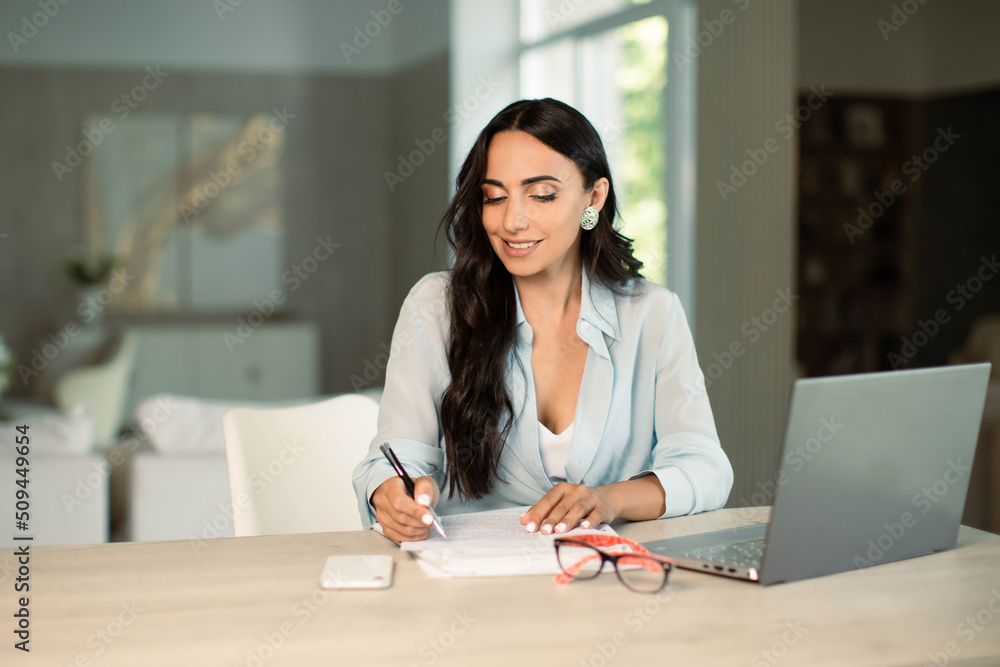 Smiling successful female using laptop computer indoors at home office, making notes on papers, remote work and small business freelancer, education and student. Smiling successful female using laptop computer indoors at home office, making notes on papers, remote work and small business freelancer, education and student.