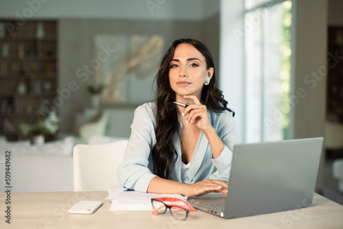 Smiling successful female using laptop computer indoors at home office, making notes on papers, remote work and small business freelancer, education and student.
