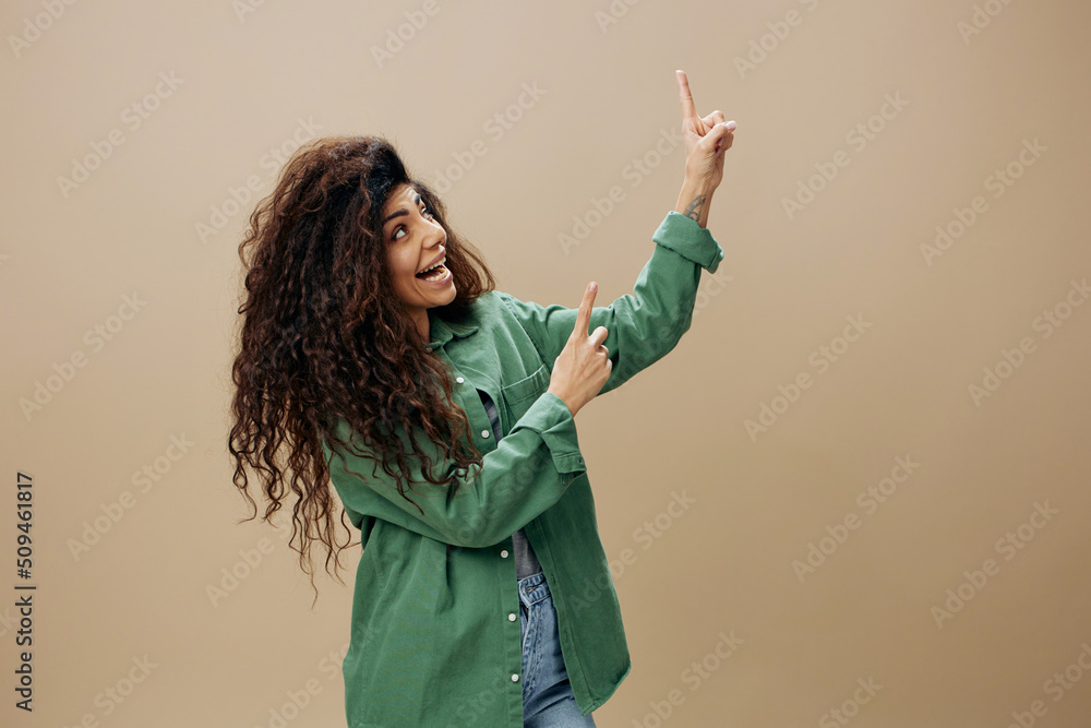 LOOK AT THIS CONCEPT. Smiling tanned curly Latin female in casual things point finger up posing isolated over pastel beige background look up show copy space Mockup Banner. Good offer LOOK AT THIS CONCEPT. Smiling tanned curly Latin female in casual things point finger up posing isolated over pastel beige background look up show copy space Mockup Banner. Good offer