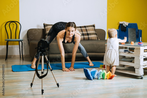 Young mother fitness instructor personal trainer record video near camera at home with baby daughter