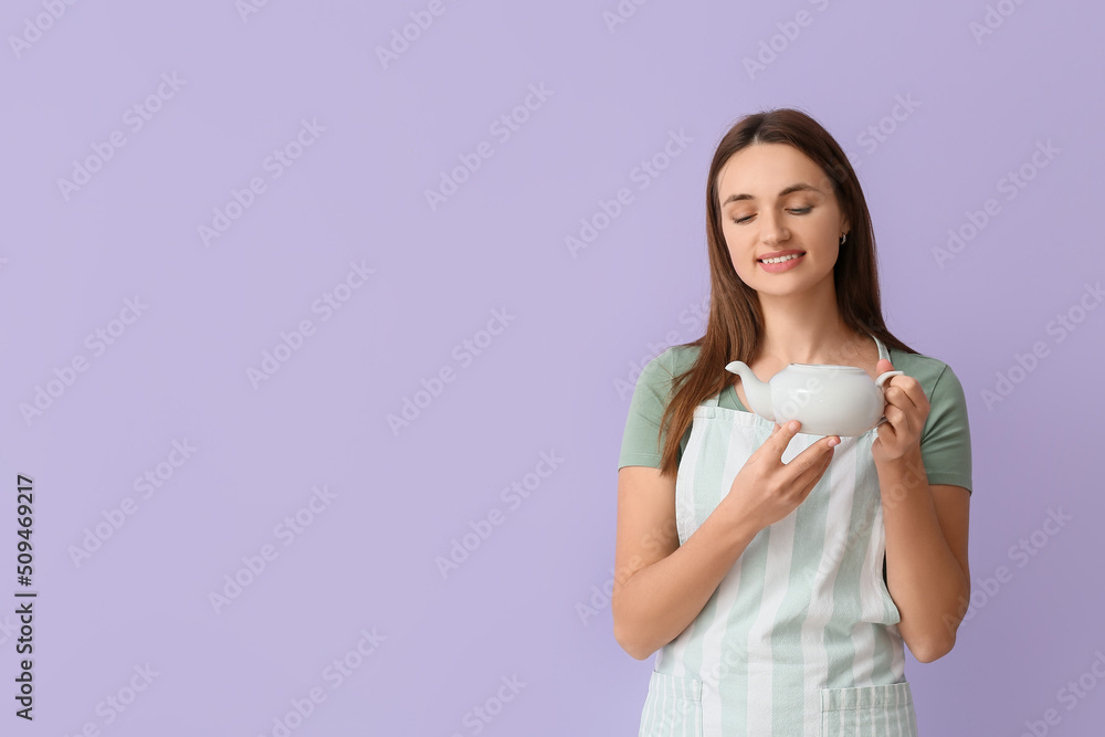 Pretty young woman in apron with teapot on lilac background Pretty young woman in apron with teapot on lilac background