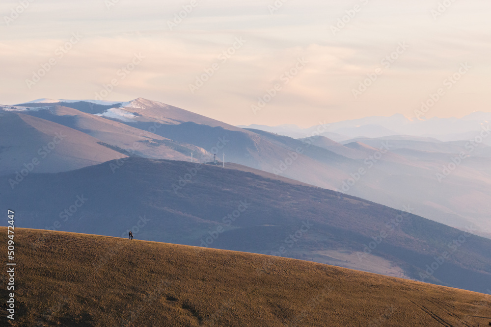 A very distant couple on top of a mountain at near sunset, with warm and soft tones and distant hills and mountains