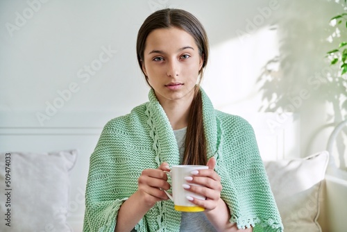 Young teenage female under knitted plaid warming up with hot tea in mug, looking at camera