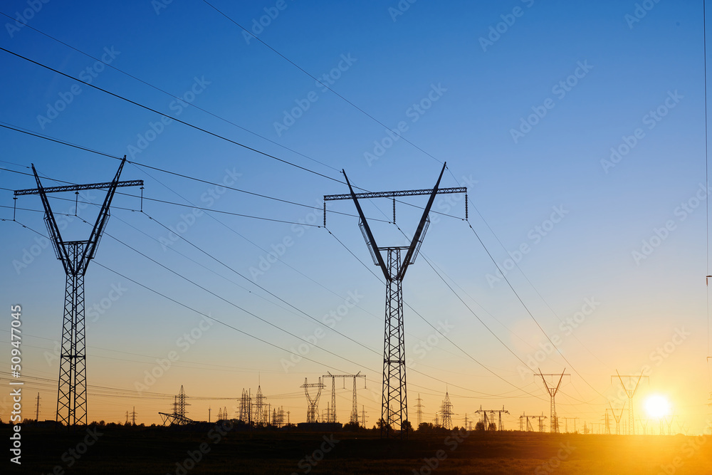 Fototapeta premium High voltage electricity towers in field at sunset and clear blue sky. Dark silhouettes of repeating power lines on orange sunrise. Electricity generation, transmission, and distribution network