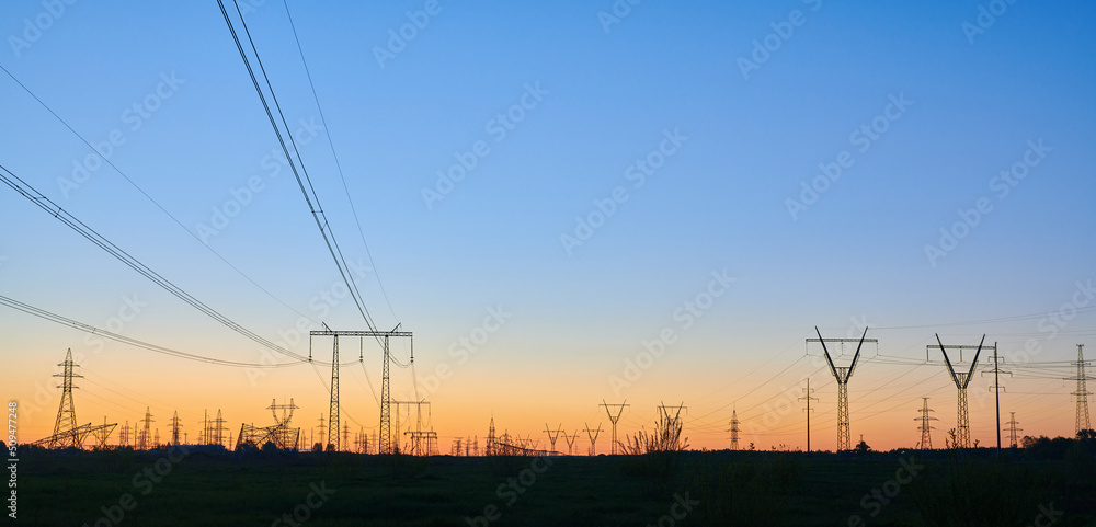 High voltage electricity towers on field at sunset and blue sky. Panoramic view of dark silhouettes of power lines on orange sunrise. Electricity generation, transmission, and distribution network