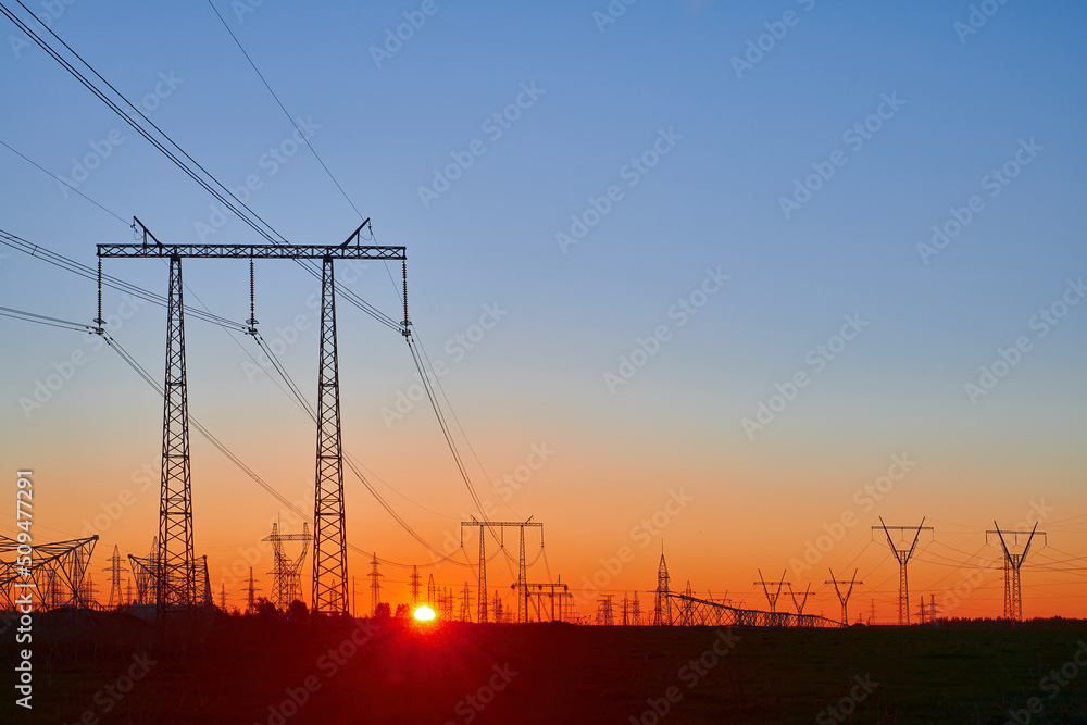 Orange sunrise on field with high voltage electricity towers. Dark silhouettes of repeating power lines on sunrise. Electricity generation, transmission, and distribution network. Industrial landscape