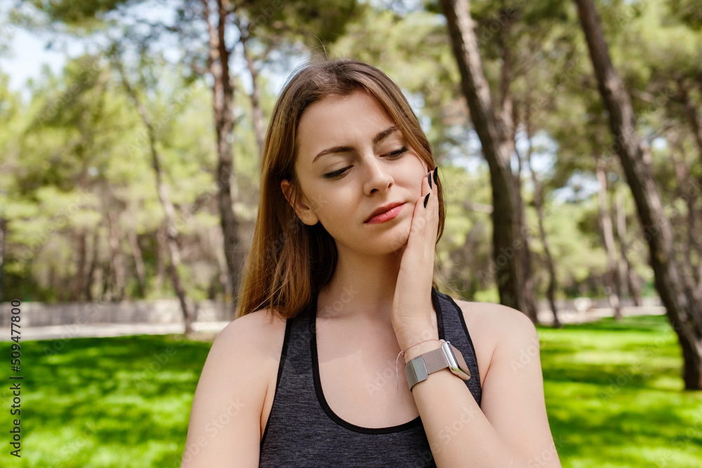 Beautiful brunette woman wearing sports bra standing on city park, outdoors touching mouth with hand with painful expression because of toothache or dental illness on teeth. Suffering from sensitive.