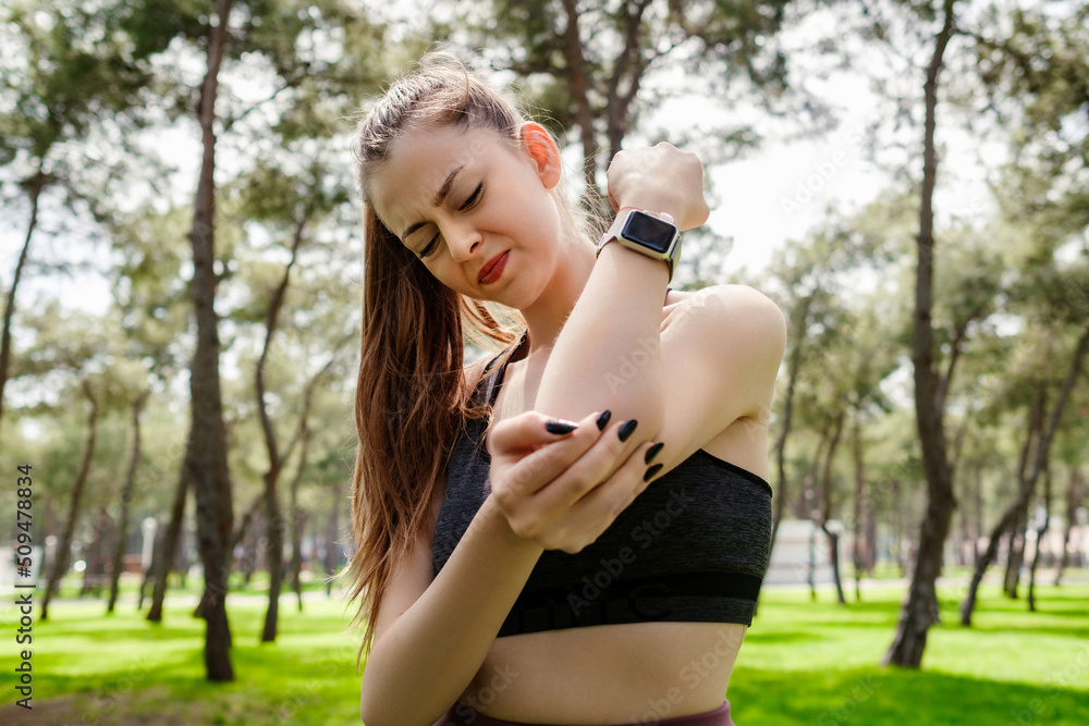 Beautiful brunette woman wearing sports bra standing on city park, outdoors suffering from chronic joint rheumatism. Elbow pain and treatment concept. Healthcare and medicine. Beautiful brunette woman wearing sports bra standing on city park, outdoors suffering from chronic joint rheumatism. Elbow pain and treatment concept. Healthcare and medicine.