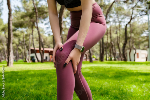 Beautiful brunette woman wearing sports bra standing on city park, outdoors touching injured knee with hands. Injured leg, healthy lifestyle and sport concept.