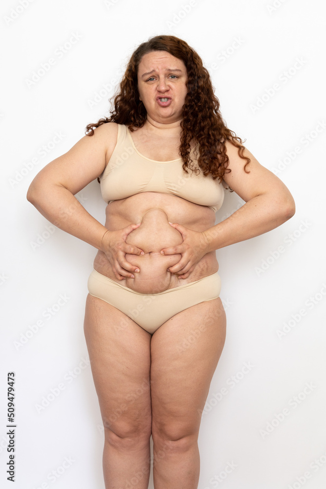 Portrait of frowning overweight woman with long curly hair wearing beige underwear, squeezing excess fat of belly, posing on white background. Weight loss, obesity, health problems, face expression. Portrait of frowning overweight woman with long curly hair wearing beige underwear, squeezing excess fat of belly, posing on white background. Weight loss, obesity, health problems, face expression.