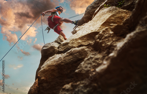 Fototapeta Muscular climber man in protective helmet abseiling from cliff rock wall using rope Belay device and climbing harness on evening sunset sky background