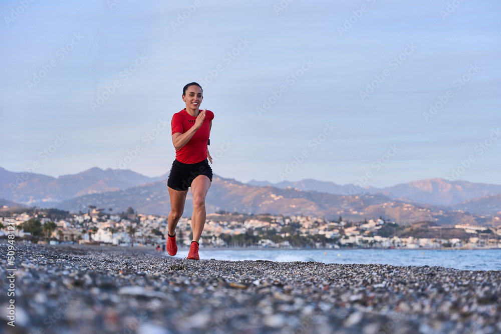 young athletic woman in a red shirt and braid running on the shore of the beach with mountains in the background young athletic woman in a red shirt and braid running on the shore of the beach with mountains in the background