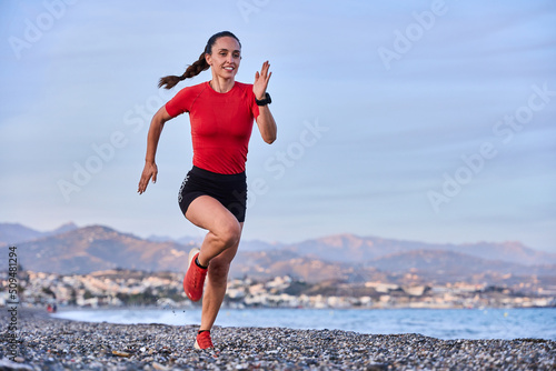 young athletic woman in a red shirt and braid running on the shore of the beach with mountains in the background
